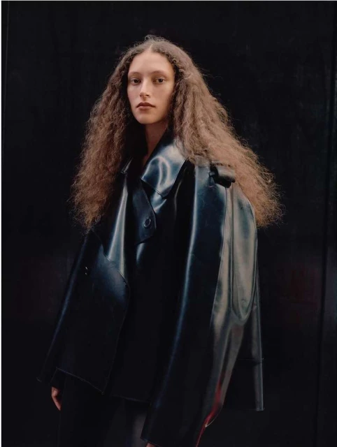 Portrait of an adult woman with long curly brown hair wearing a shiny black vinyl coat posing against a dark background in a photo studio.