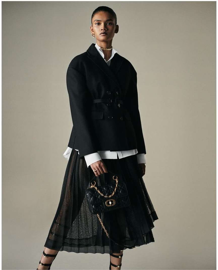 Fashionable woman in a black jacket and pleated skirt holding a black quilted handbag with gold chain, studio shot.