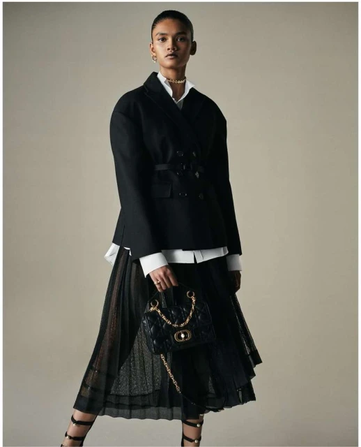 Fashionable woman in a black jacket and pleated skirt holding a black quilted handbag with gold chain, studio shot.
