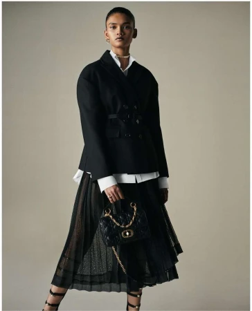 Fashionable woman in a black jacket and pleated skirt holding a black quilted handbag with gold chain, studio shot.