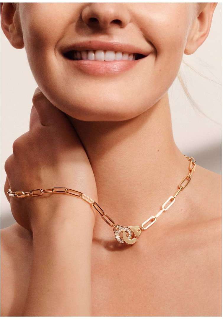 Close-up of a fair-skinned woman with a gold chain link necklace featuring a handcuff pendant, set against a cream-colored backdrop.