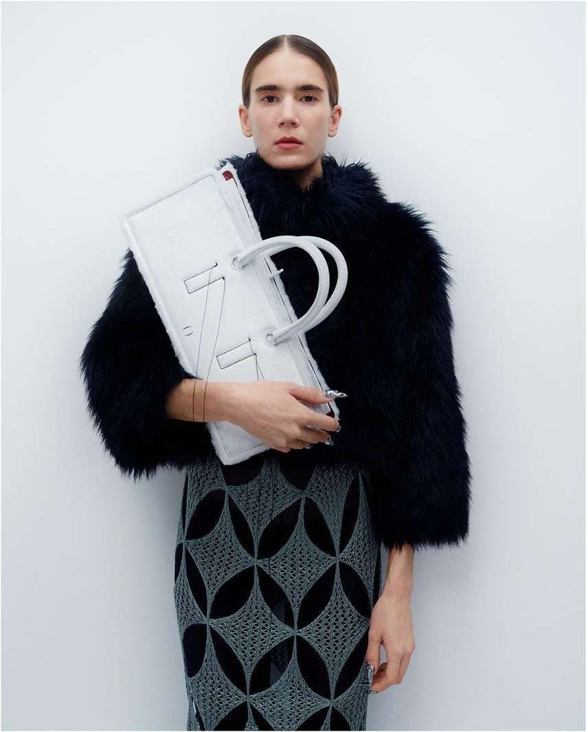 Fashionable woman with dark faux-fur jacket holds a large white handbag with graphic stitching, wearing a patterned pencil skirt.
