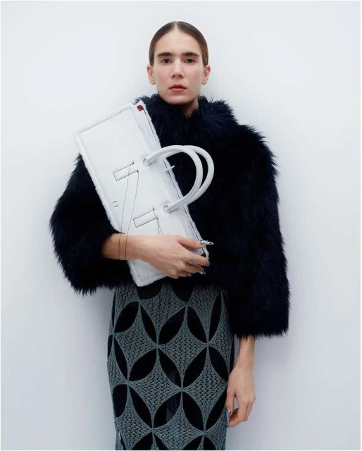 Fashionable woman with dark faux-fur jacket holds a large white handbag with graphic stitching, wearing a patterned pencil skirt.