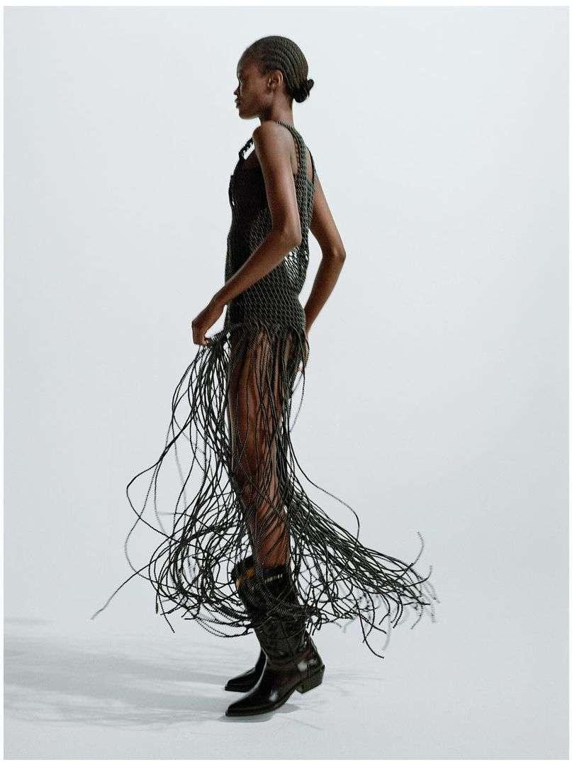 Full-length shot of an African American woman in a dress with fringe, wearing cowboy boots against a bright neutral backdrop in studio.