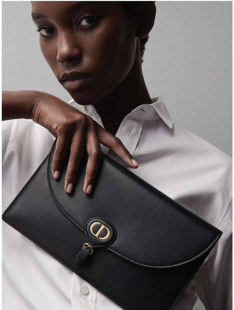 Elegant African American woman showcases a black leather Dior clutch purse, emphasizing fashion and luxury against a neutral backdrop.
