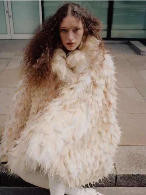 Fashionable woman with voluminous curly hair poses outside wearing a large off-white feathery textured coat and light-colored pants and boots.