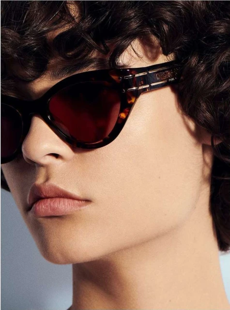 Close-up portrait of a woman with curly hair wearing brown tortoiseshell sunglasses in soft lighting, focusing on her face and stylish eyewear.