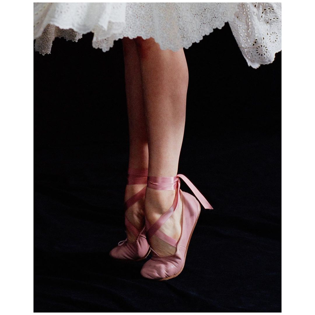 Ballerina's feet in pink pointe shoes and ribbons, standing with white tutu against a dark background in an indoor setting.