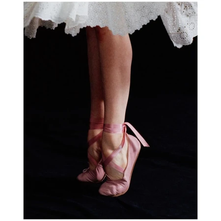 Ballerina's feet in pink pointe shoes and ribbons, standing with white tutu against a dark background in an indoor setting.