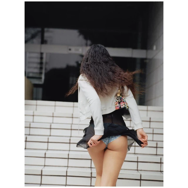 A young adult female walks up outdoor steps. She is wearing a light jacket, short black skirt, and a small colorful handbag.