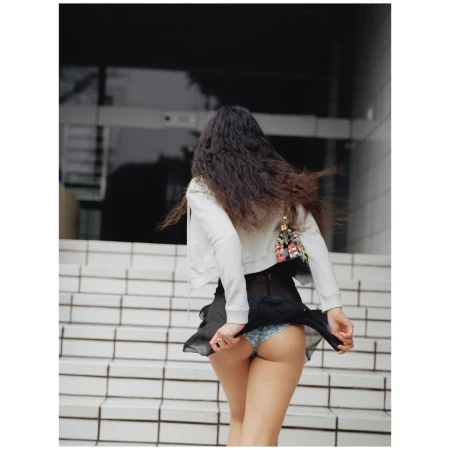 A young adult female walks up outdoor steps. She is wearing a light jacket, short black skirt, and a small colorful handbag.
