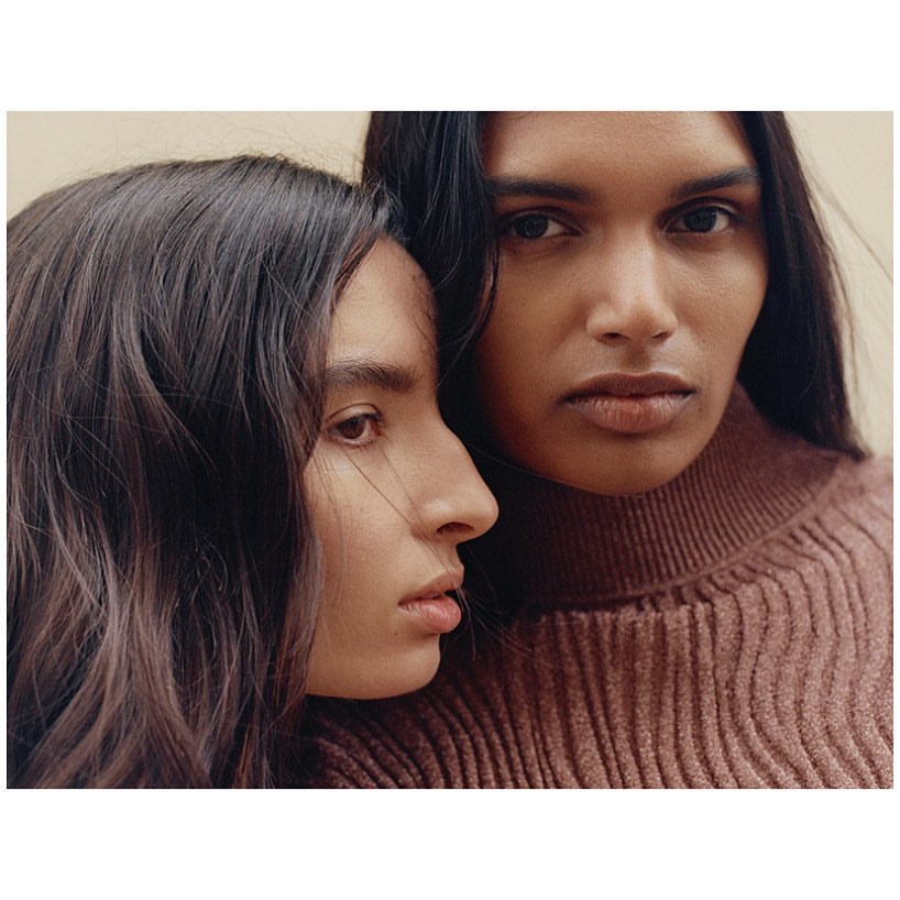 Two women pose indoors, close-up. One woman looks directly at the camera wearing a brown turtleneck sweater, indoor setting, neutral background.