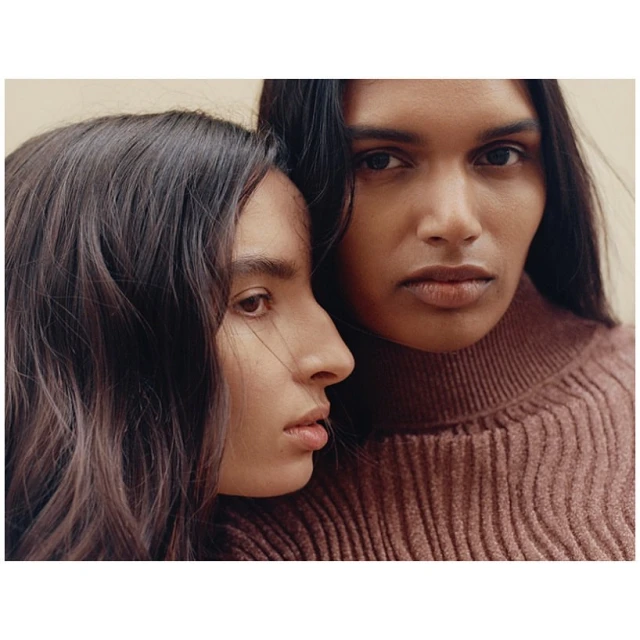 Two women pose indoors, close-up. One woman looks directly at the camera wearing a brown turtleneck sweater, indoor setting, neutral background.