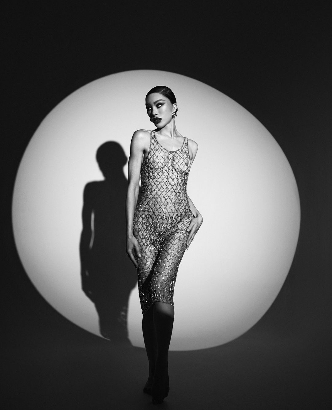Elegant woman in a sparkling dress posing in front of a round light, captured in black and white for a high-fashion aesthetic.