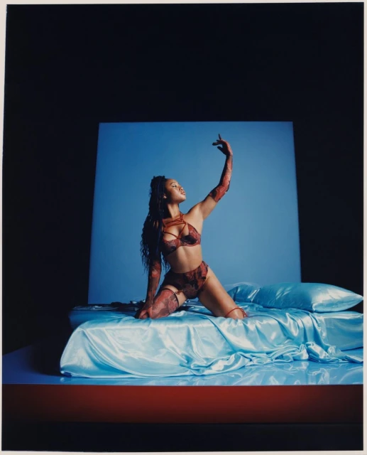 Adult female model poses on a bed with a blue sheet against a solid blue backdrop wearing a red lingerie set in a photo studio.