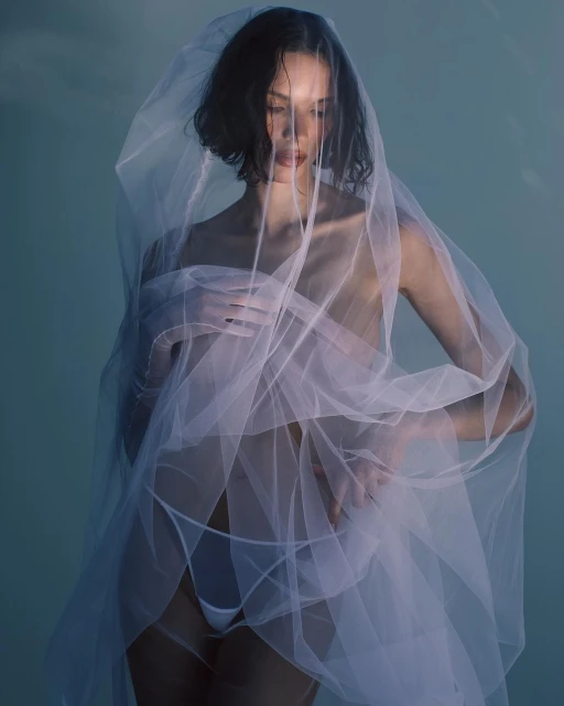 Woman with short dark hair poses indoors, draped in white translucent fabric against a light blue backdrop in a fashion-style portrait.