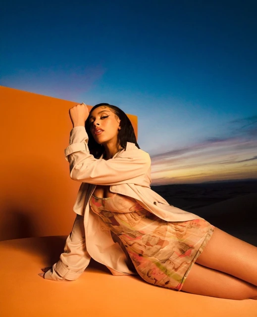 Portrait of a stylish woman in a floral dress and beige coat sitting against an orange backdrop with a gradient sky background.