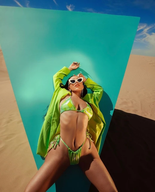 Woman in lime-green bikini and jacket poses against turquoise backdrop in desert with blue sky. White sunglasses, long dark hair.