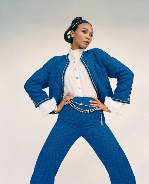 Zoe Saldana poses in a bright blue Chanel suit with gold accents and pearl accessories against a neutral backdrop in a studio shot.