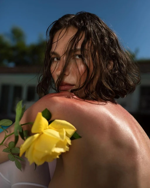 Brunette woman with damp hair poses near a pool, holding a yellow rose, with a soft focus backdrop of trees and a bright blue sky.