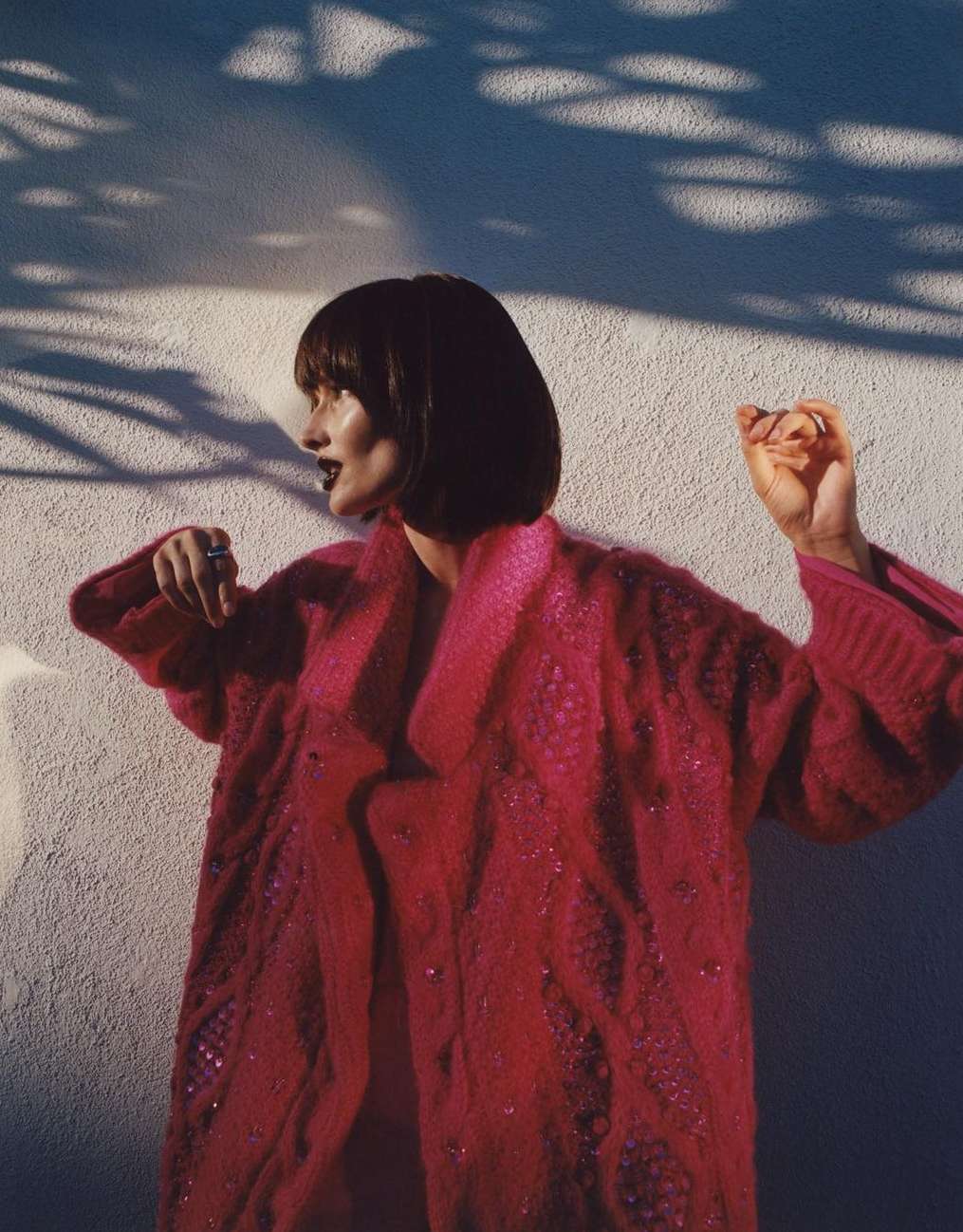 Woman in a vibrant pink bouclé coat with dark lipstick poses in front of a sunlit wall with striking leaf shadows creating a dynamic backdrop.