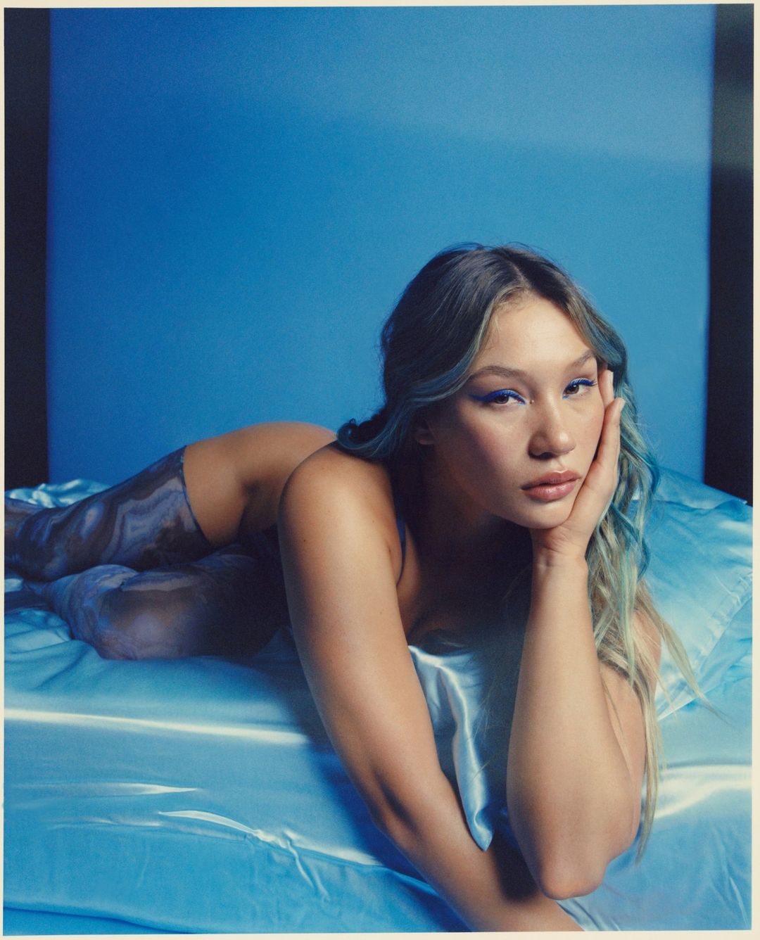 Woman with blue eyeshadow lying on a satin bed with a blue backdrop in a studio, looking at the camera with hand on cheek.