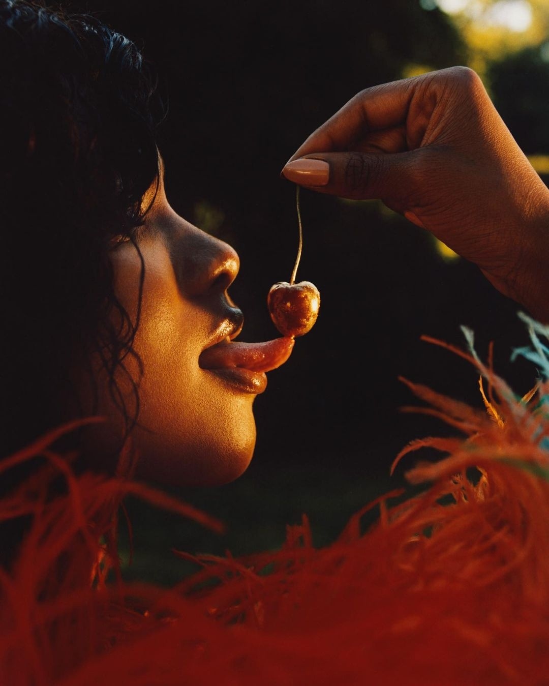 A side profile of an adult woman licking a cherry held by a hand against a dark background with orange feather details at the bottom.