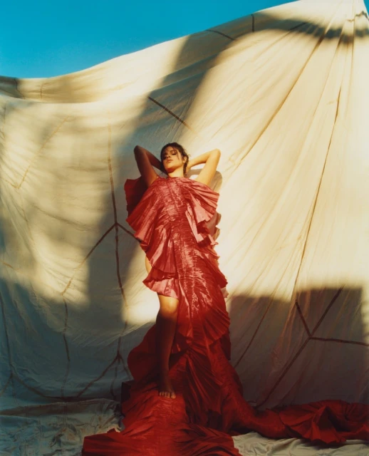 A woman in a red ruffled dress poses in front of a tan cloth, her arms raised, bare feet visible, and clear blue sky above.