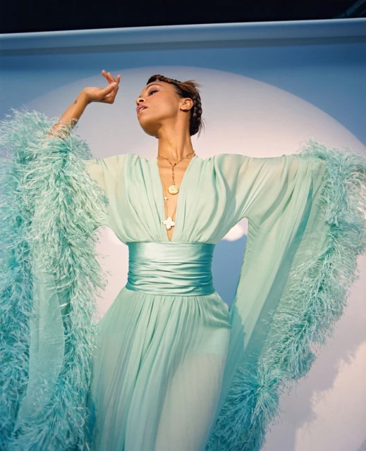 Elegant woman in flowing turquoise gown with feather trim, posing against a pale blue backdrop with soft, diffused light.