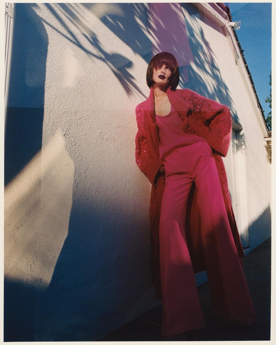 Stylish woman in a pink monochrome ensemble poses against a textured wall, accentuated by striking leaf shadows and a sunny ambiance.
