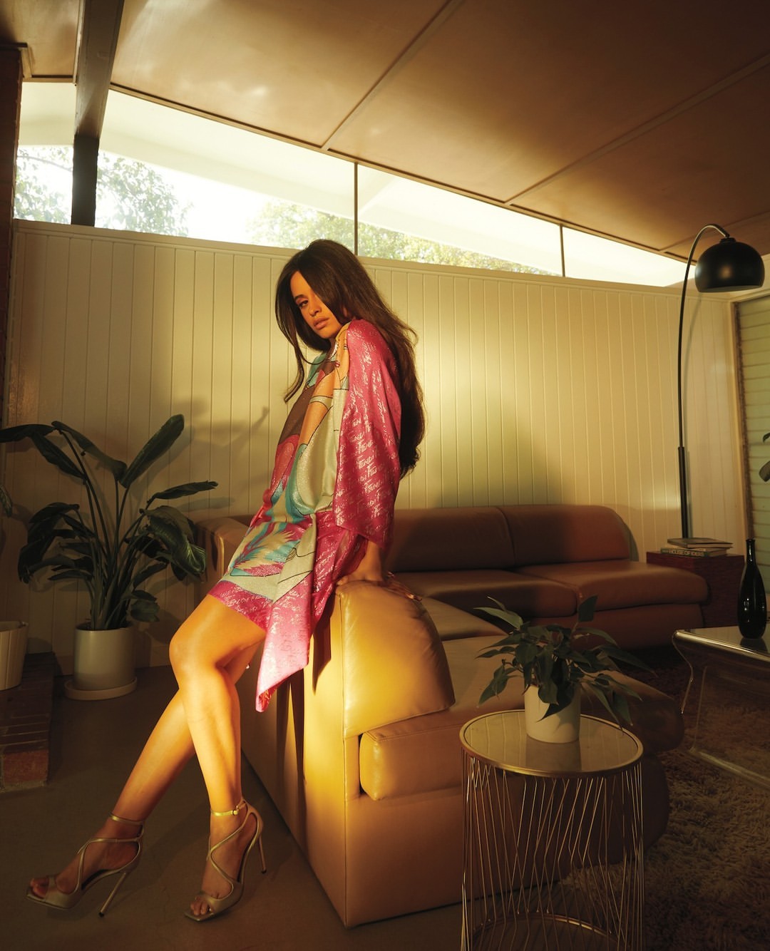 Brunette woman in pink dress posing on leather couch in modern living room with plants and decorative lighting, shot at eye-level.