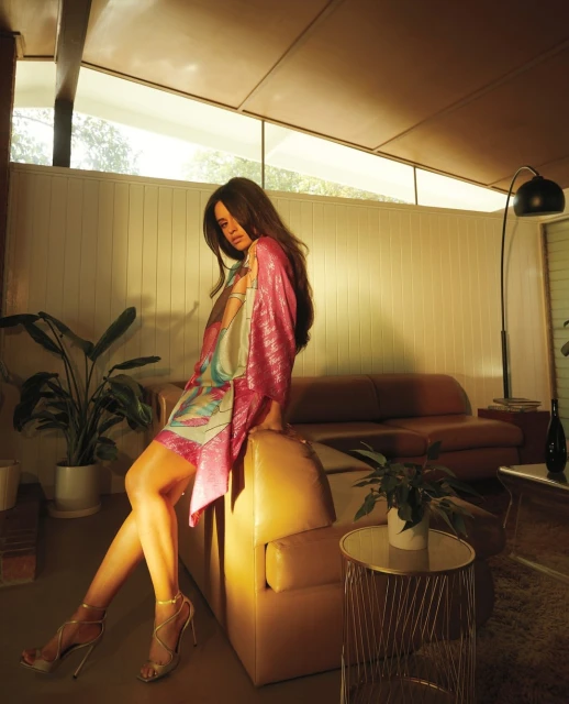 Brunette woman in pink dress posing on leather couch in modern living room with plants and decorative lighting, shot at eye-level.