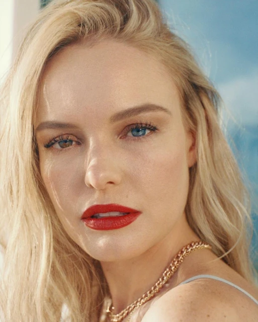 Close-up beauty portrait of a blonde woman with heterochromia wearing red lipstick and a gold chain necklace against a blue backdrop.