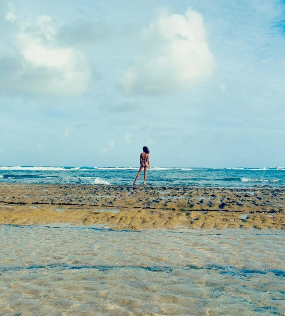 A woman in swimwear standing on a rock formation by the ocean with blue water and sky filled with white clouds during the daytime.