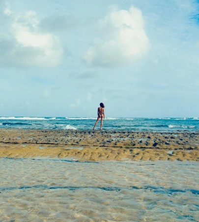A woman in swimwear standing on a rock formation by the ocean with blue water and sky filled with white clouds during the daytime.