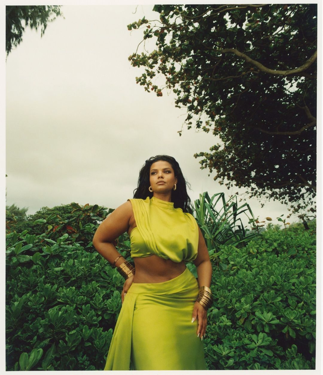 A woman in a flowing green dress poses in a lush outdoor setting, with greenery surrounding her and an overcast sky above.