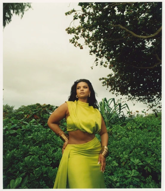 A woman in a flowing green dress poses in a lush outdoor setting, with greenery surrounding her and an overcast sky above.
