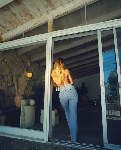 Adult woman in jeans poses in a doorway with blonde hair. Interior visible through glass with stone wall and wood-beam ceiling.