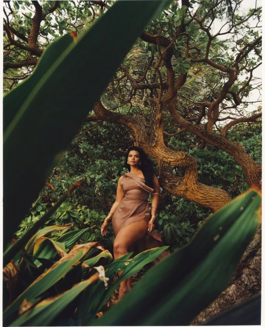 An adult woman poses in a sheer brown dress, surrounded by lush green foliage, with a large tree in the background, creating a natural, ethereal setting.