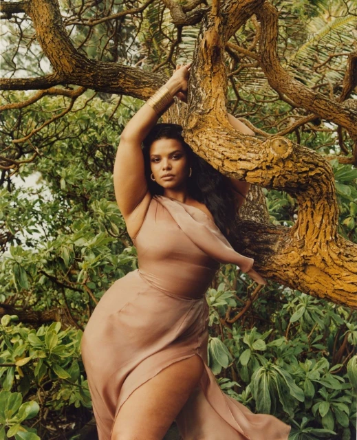 Woman in flowing dress poses near a tree in a natural outdoor setting with lush green foliage and dappled sunlight.