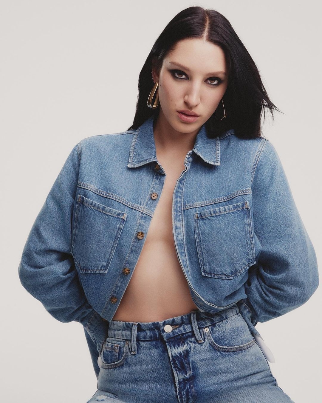 Woman in denim jacket and jeans poses in studio. The jacket is unbuttoned, revealing her chest. She wears gold hoop earrings.