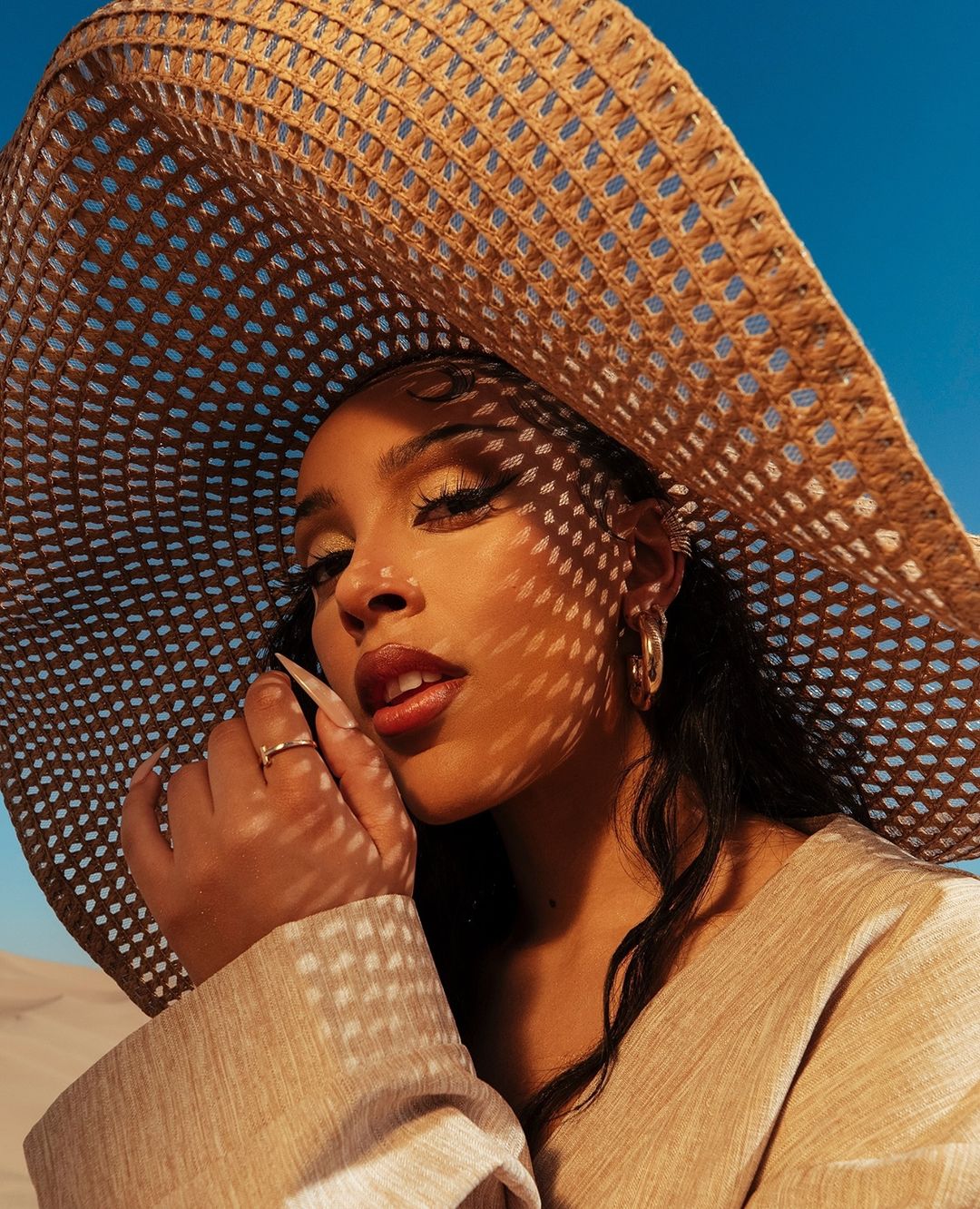 Young woman in a large straw sunhat poses in a beige outfit, casting a shadow over her face in a desert environment under a blue sky.