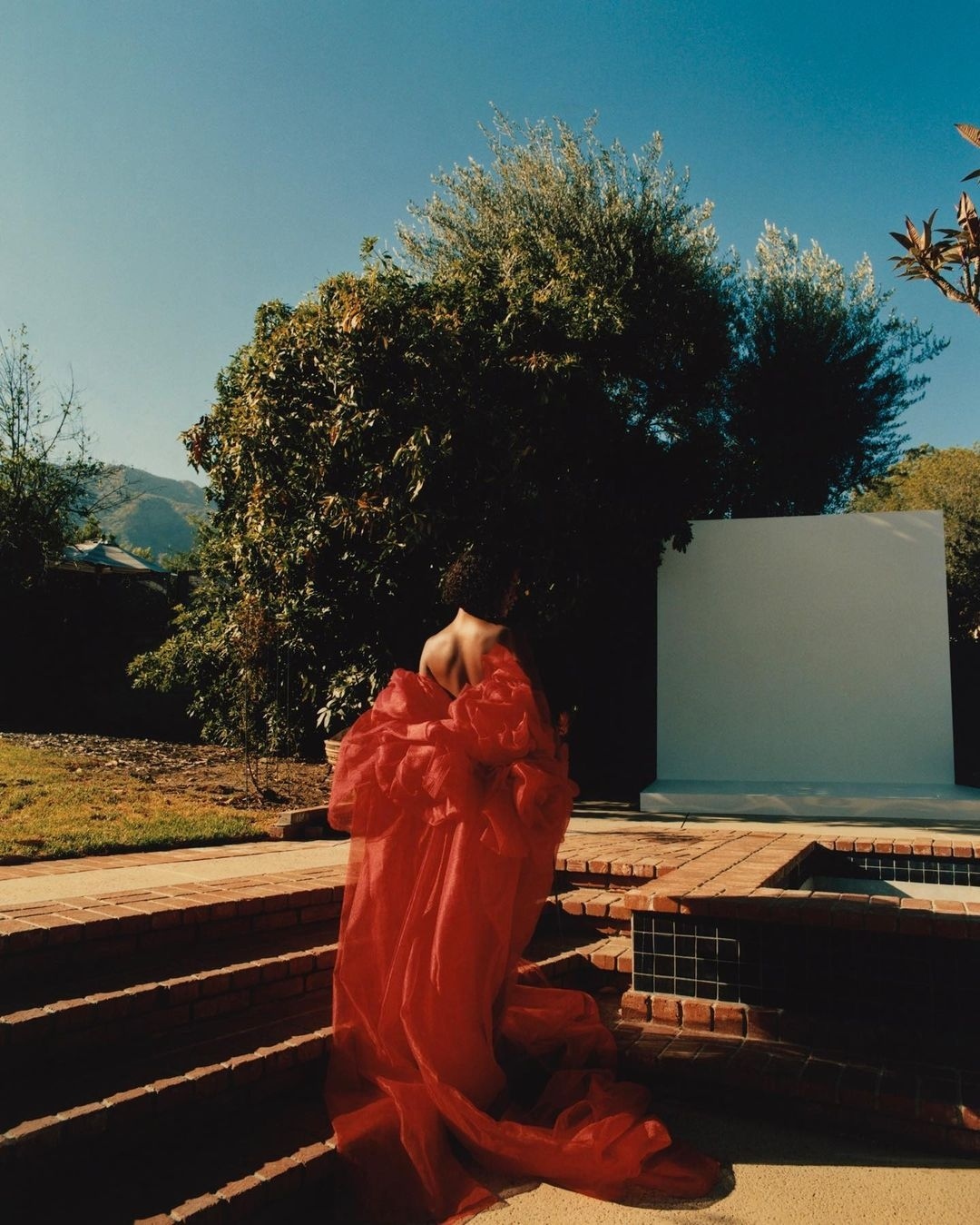 Woman wearing red couture gown, walking on terracotta steps in a garden with trees and mountains in the background under blue skies.