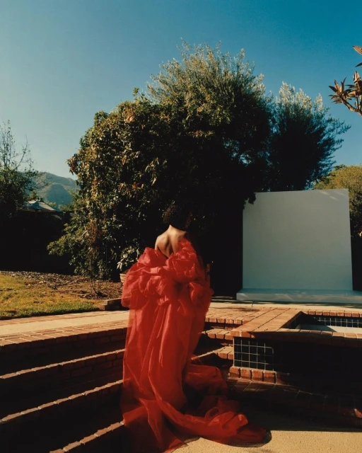 Woman wearing red couture gown, walking on terracotta steps in a garden with trees and mountains in the background under blue skies.