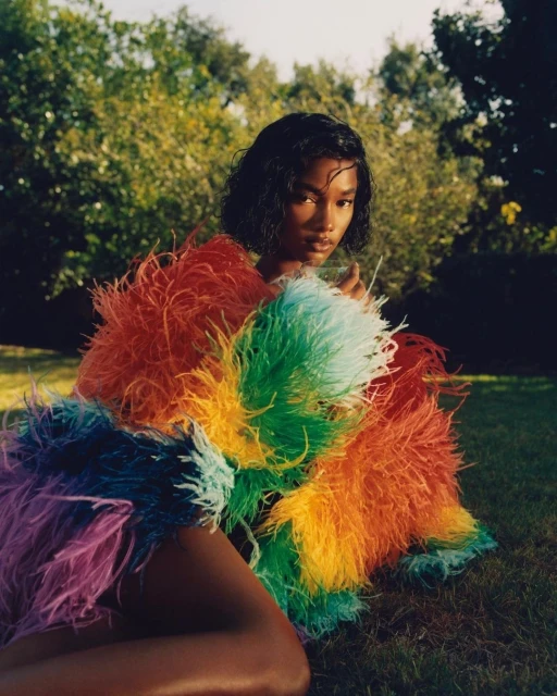 An adult female is posed in a field wearing a multi-colored, feathery garment; trees and grass are visible in the background.