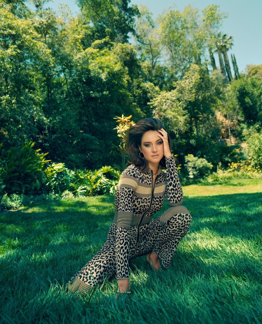 A woman in a leopard-print outfit poses in a lush green meadow surrounded by trees on a sunny day, looking directly at the camera.