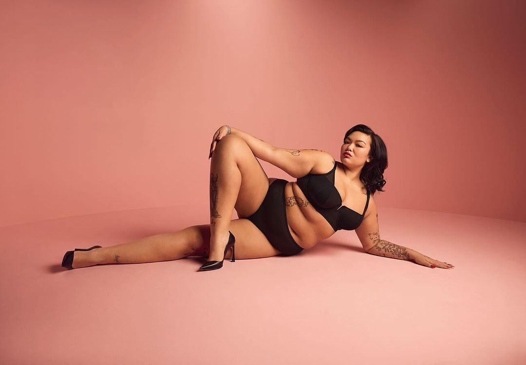 Plus-size woman lying down in black lingerie and heels in a studio setting with soft pink backdrop, showcasing body positivity and fashion.
