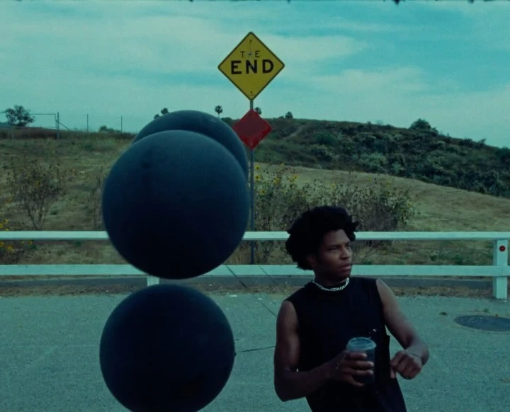 Young adult male in black sleeveless shirt holding a cup, standing on a road next to large black spheres, under a 'The End' sign.