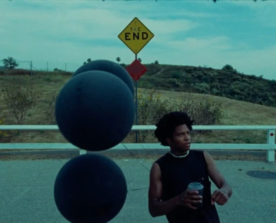 Young adult male in black sleeveless shirt holding a cup, standing on a road next to large black spheres, under a 'The End' sign.