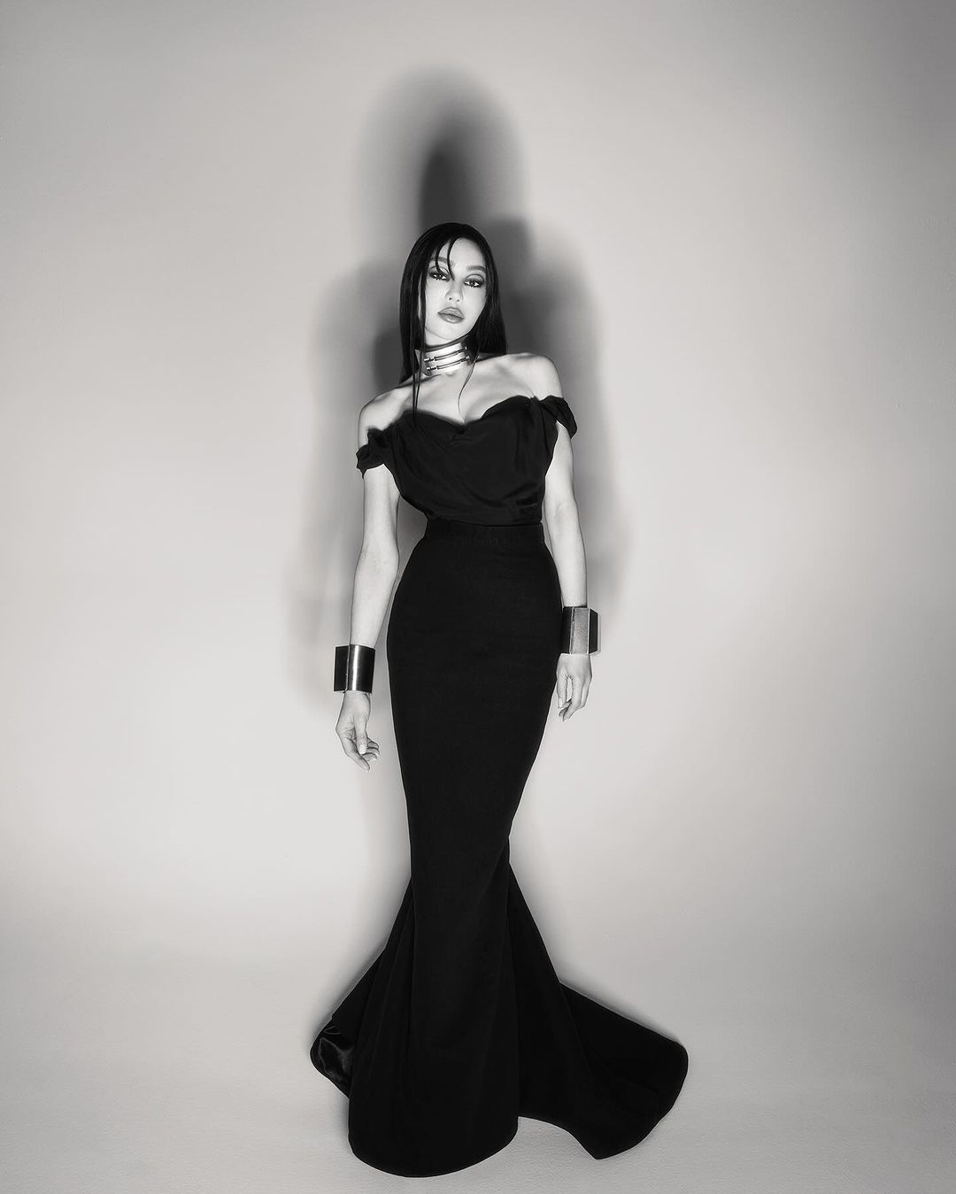 Woman in black off-the-shoulder dress with silver choker and bracelets poses in a photo studio. Black and white fashion shot.
